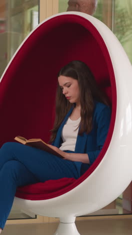 woman reads book in egg chair in library. concentrated brunette lady rests with old female novel in comfortable armchair. contemporary reading hall