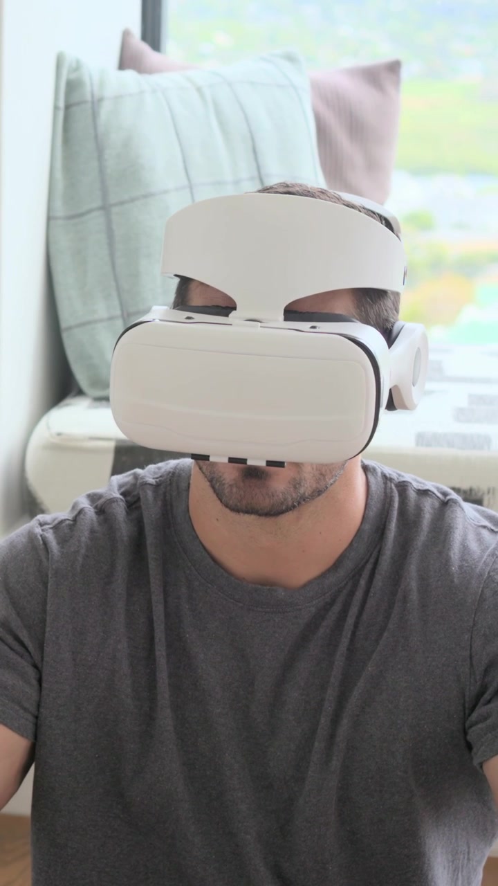 Video verticale: Uomo in maglietta grigia che si allunga, si sistema un visore VR bianco a casa con i cuscini, esplorando.