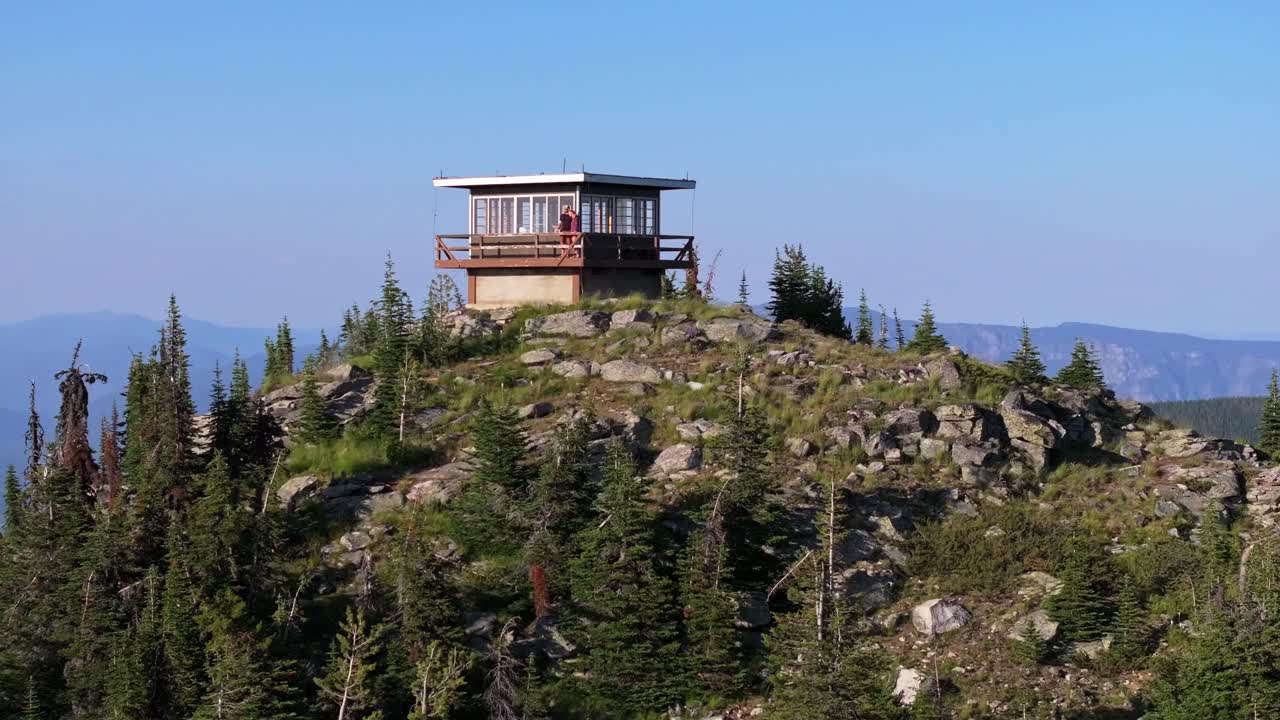 En droneoptagelse, der trækker sig væk fra et par, der står på verandaen i et isoleret brandtårn på toppen af et bjerg i det fjerne nordlige Idaho foran et fantastisk, robust landskab