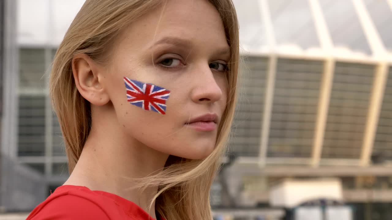 портрет молодой блондинки, футбольной фанаты из англии, смотрящей в камеру, серьезное лицо, стадион на заднем плане, 50 кадров в секунду.
