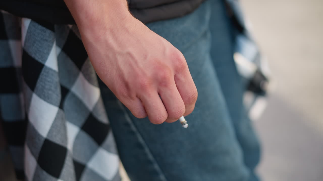 close up of man's hand holding cigarette attempting to flick off ash that stays intact, wearing denim jeans and checkered shirt tied at waist with soft background blur and outdoor lighting