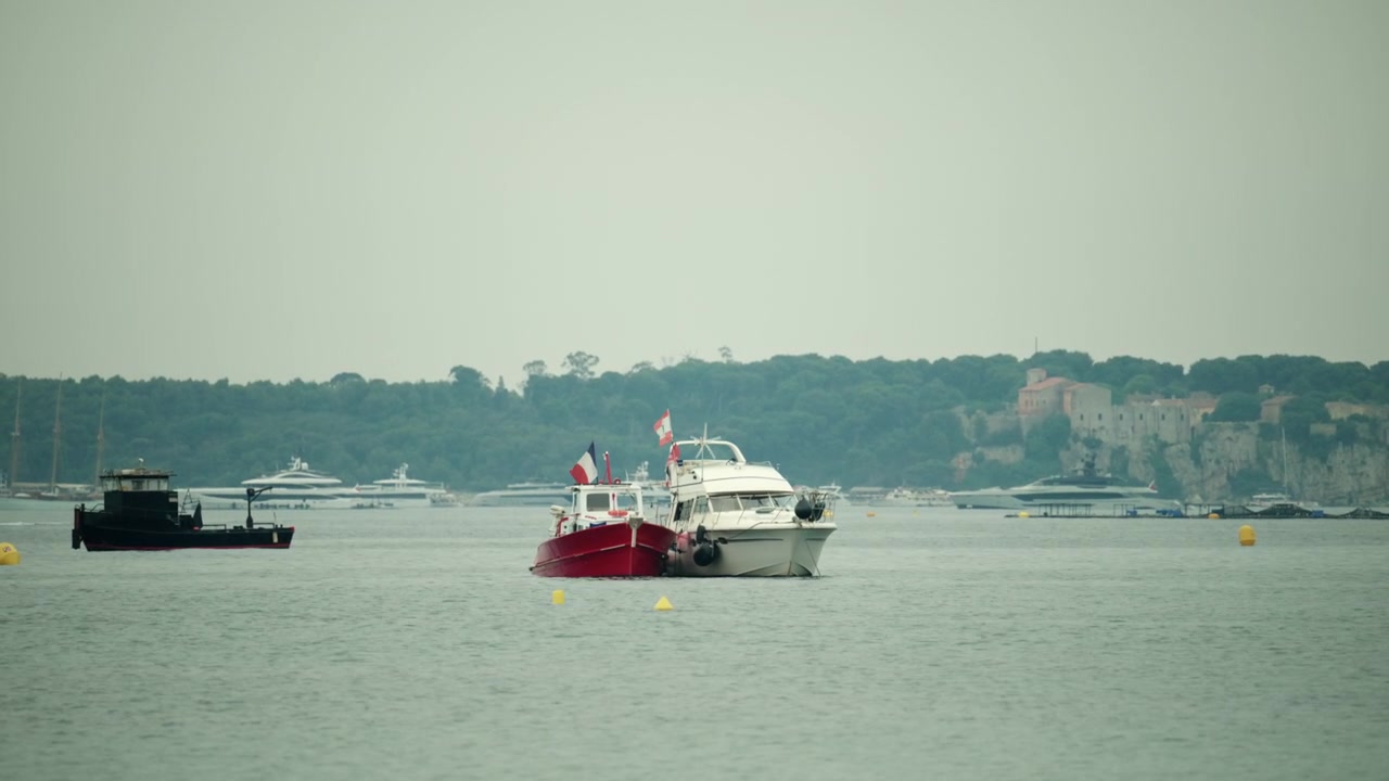Barcos navegando pelas águas da Riviera Francesa, com uma cidade costeira e colinas ao fundo. Bandeiras tremulando em duas embarcações.
