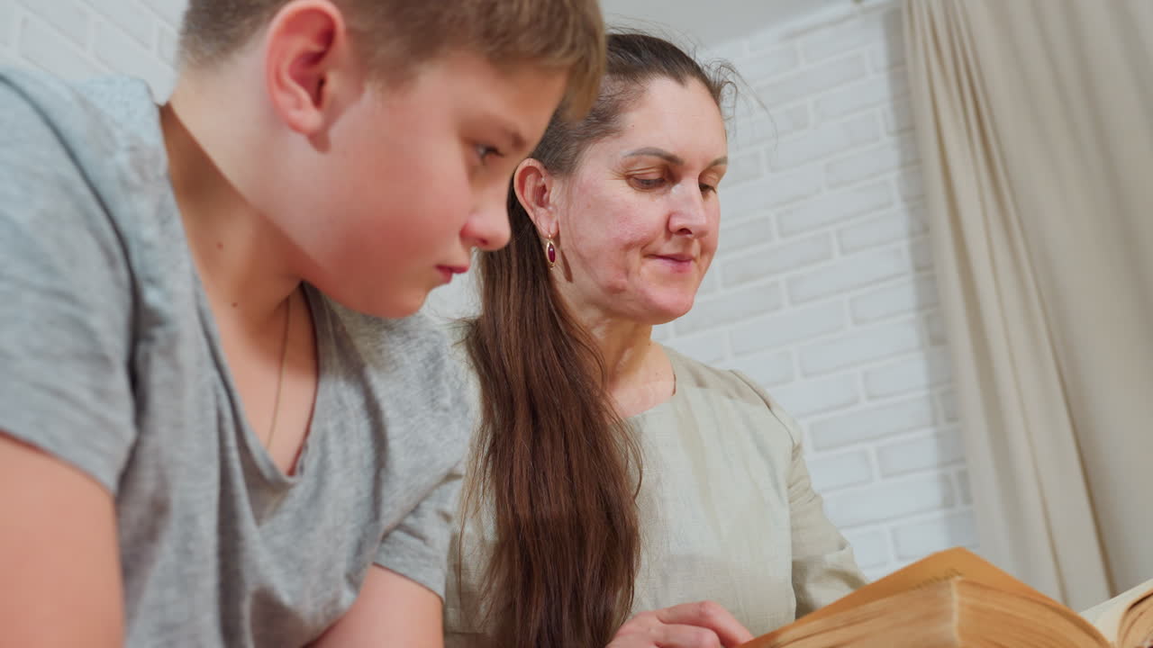 close view of woman in beige gown and boy in grey shirt sitting closely while attentively looking into old thick book during relaxed indoor moment with white brick wall and beige curtain