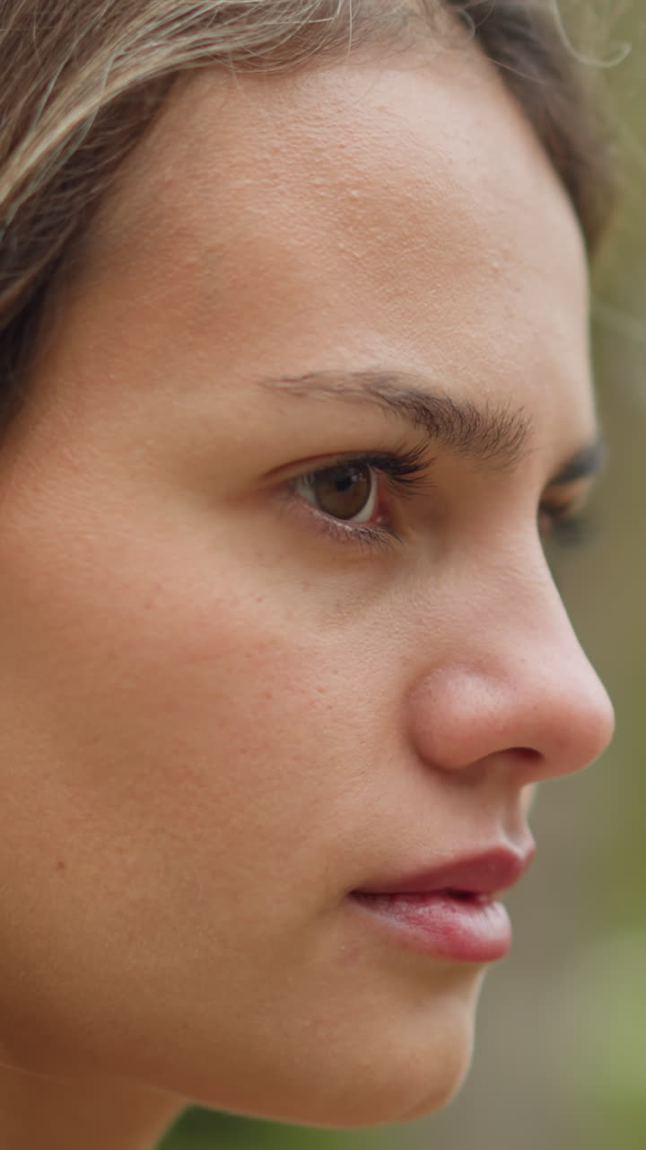 Close-up view of womans face turning and blinking eyes, subtle expression, natural beauty, soft focus, relaxed moment, outdoor setting, calm and serene expression, slow motion capture