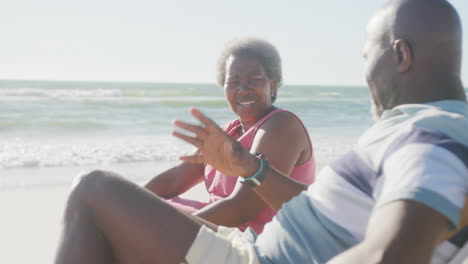 happy senior african american couple sitting on deck chairs and talking at beach, in slow motion