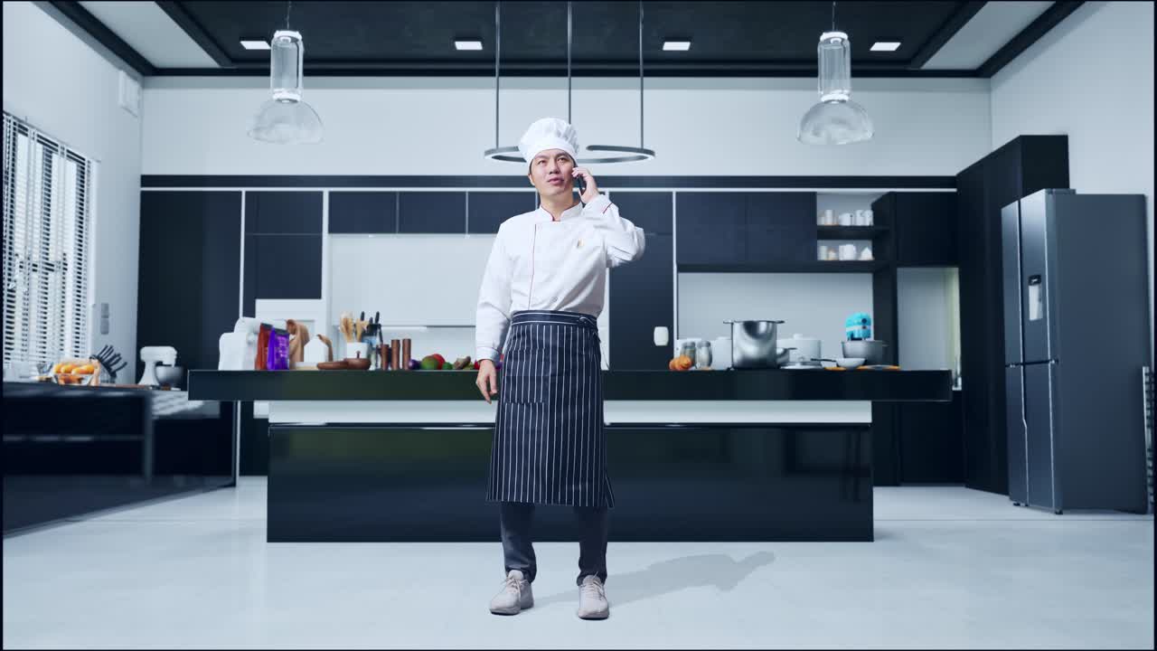 Figura intera di chef asiatico che parla al telefono e si guarda intorno mentre è in piedi nella cucina di casa