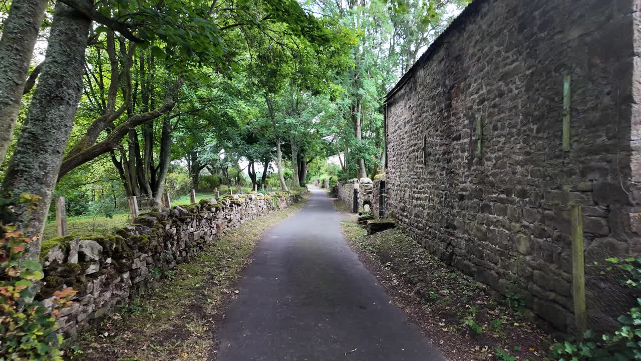 Paved road going through the english countryside with stone walls, trees and a stone building
