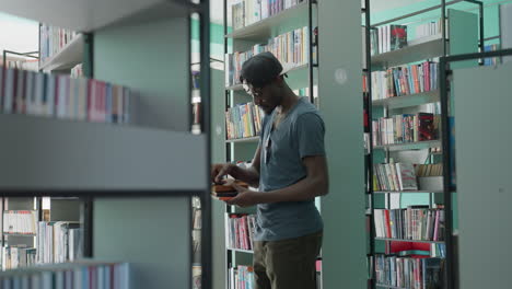 Young man in casual attire and cap browsing library shelves, holding book with red cover while flipping through pages, surrounded by organized rows of colorful books