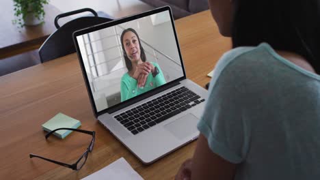mujer de negocios de raza mixta sentada en un escritorio usando una computadora portátil haciendo una videollamada con una colega
