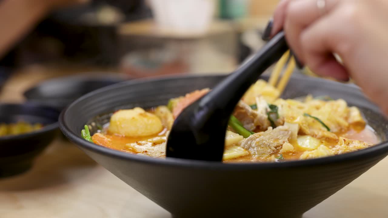 Hand stirs hearty Asian hotpot with tofu, meat, vegetables in savory chili broth, close-up