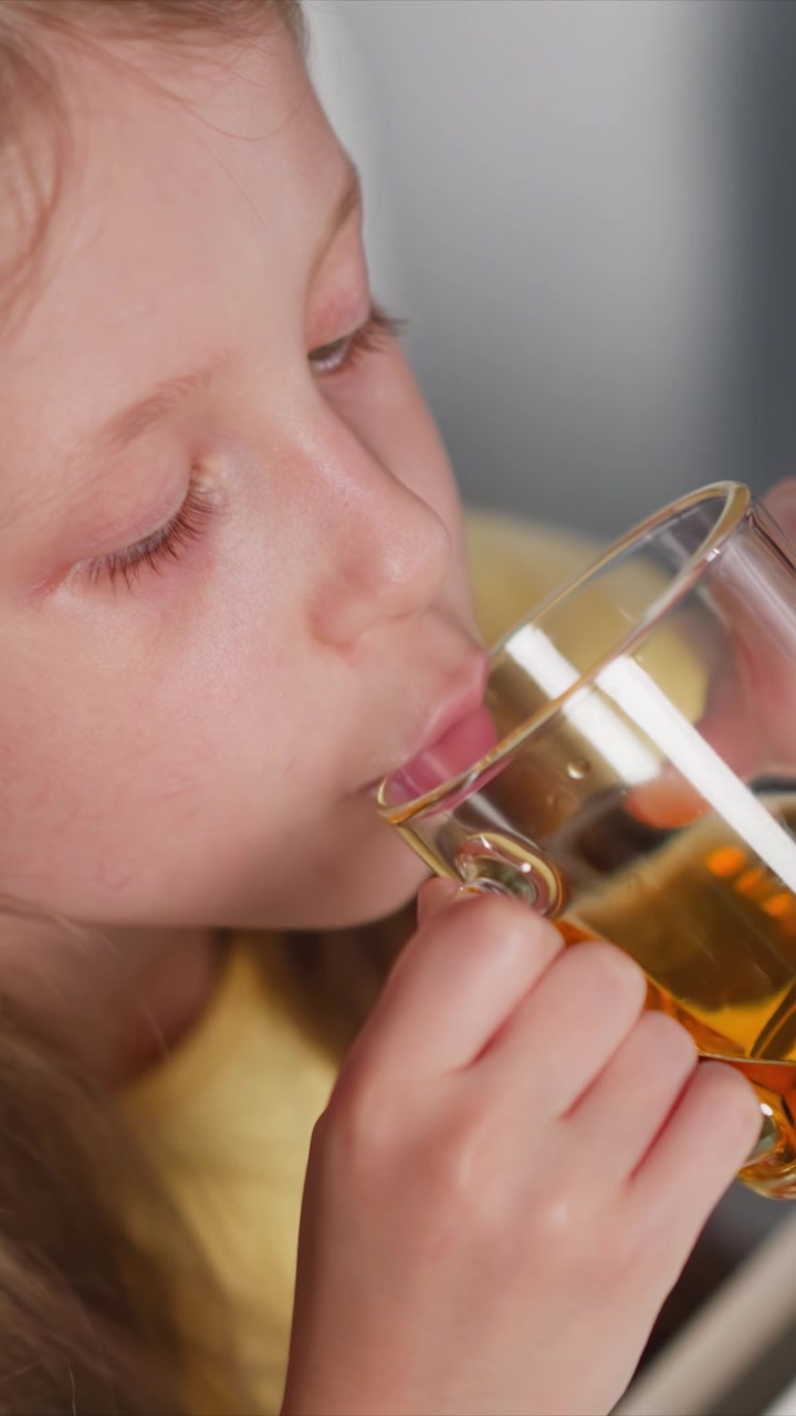 Primo piano di una bambina che beve da un bicchiere con liquido dorato. La bambina tiene una tazza trasparente vicino alla bocca, concentrandosi sull'attività di bere. L'illuminazione soffusa evidenzia i dettagli negli scatti ravvicinati.