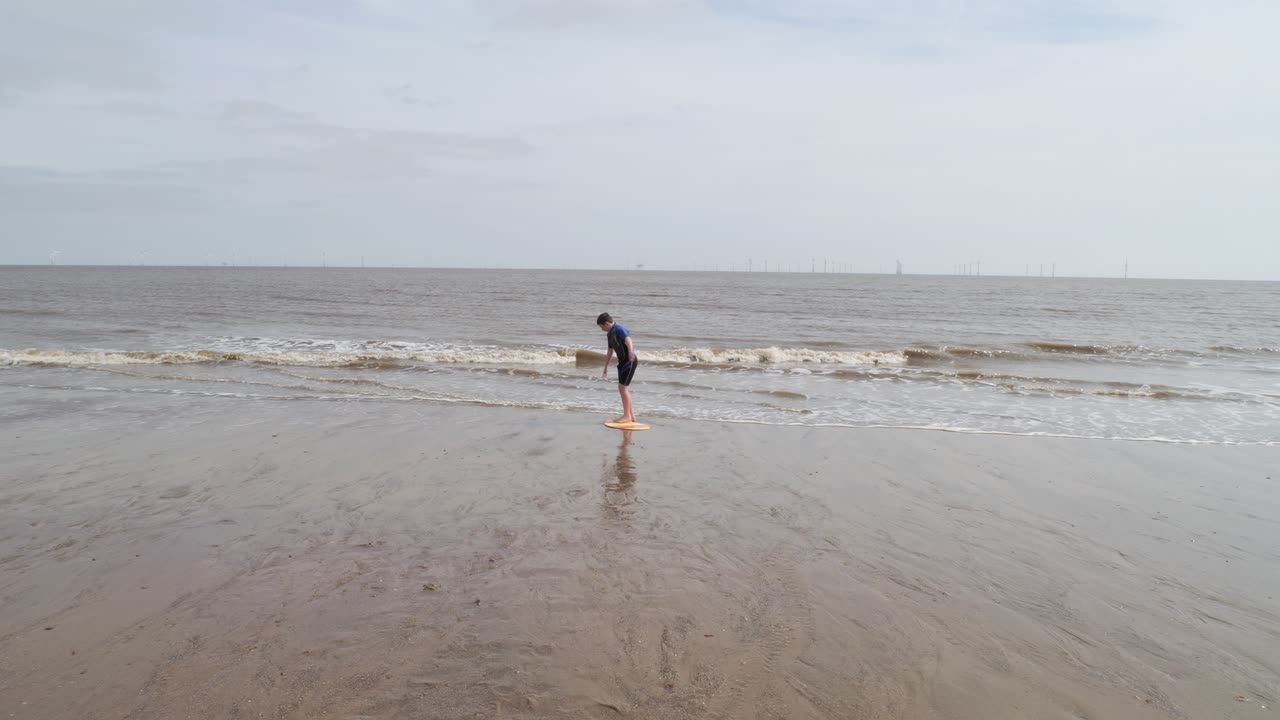 Young boy in a wetsuit on a beach digging in the sand. Playing with a surfboard and practicing skimming on the water. Seaside scene. Ocean waves breaking on the beach. Holiday time season