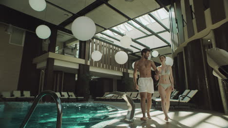 cheerful couple walking indoor pool. happy couple holding hands poolside