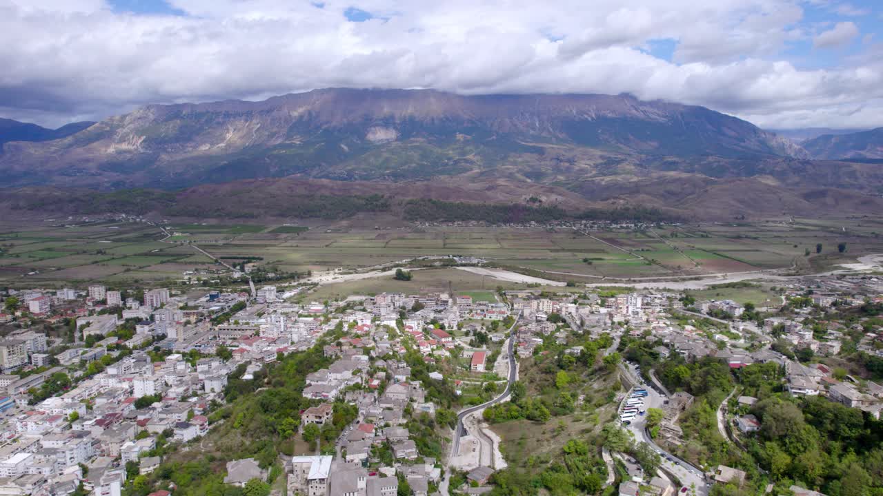 Ampia ripresa aerea con drone di una piccola città con strade tortuose e terreni agricoli circostanti, sullo sfondo di una grande catena montuosa sotto nuvole spettacolari, movimento fluido della camera con panoramica