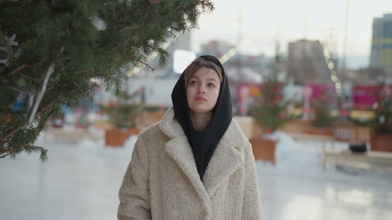 niña con una cálida chaqueta de invierno beige y una bufanda con capucha negra camina por una calle festiva nevada, mirando a un árbol verde bellamente decorado. detrás de ella, casas borrosas y una luz de cuerda brillante