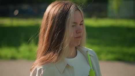 young woman stands in bright sunlight, cooling her face with a green air pump, squinting slightly as gentle breeze flows through her hair, sunlight filters through trees and a building in background