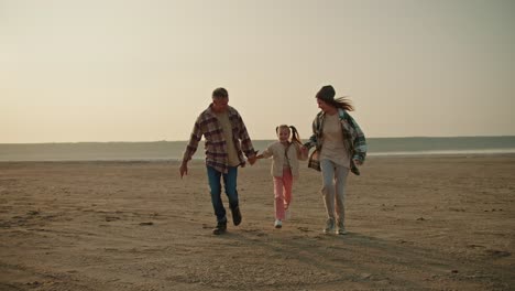 A-happy-family,-a-middle-aged-brunette-man-in-a-checkered-shirt,-together-with-his-wife,-a-brunette-girl-in-a-green-checkered-shirt,-are-running-along-a-deserted-seashore-and-lifting-up-their-little-daughter,-a-blonde-girl-in-a-white-jacket-and-pink-pants,-during-their-vacation-outside-the-city