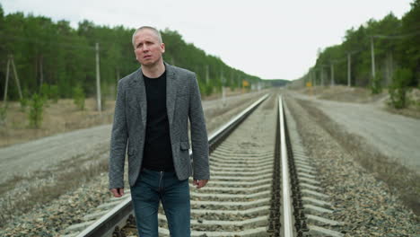 a close-up view of a middle-aged man wearing a grey suit jacket and black shirt, walking along railway tracks in a forested area