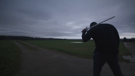 Modern-Black-Swordsman-Warrior-Awaiting-Battle-Opponent-in-Open-Field-Under-Grey-Sky,-Europe,-4K-|-Muscular,-Intimidating,-Shadow,-Figure,-Blurry,-Dark-Clothes,-Powerful,-Adjusting-Stance,-Patrol