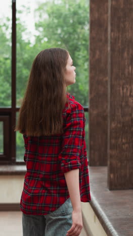 woman looks through terrace window. tranquil lady with loose red hair wearing casual clothes enjoys panoramic view from studio apartment. cosy loneliness