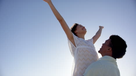 Biracial-couple-enjoys-a-sunny-day-outdoors,-woman-reaching-towards-the-sky