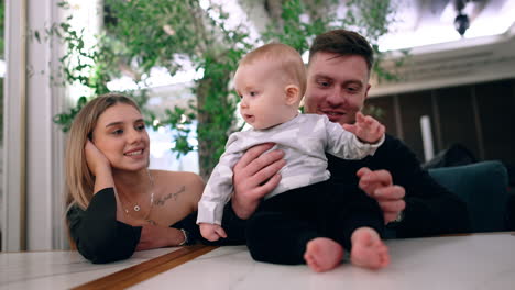loving caucasian parents sitting at table. lovely cute baby sits on the table supported by dad.