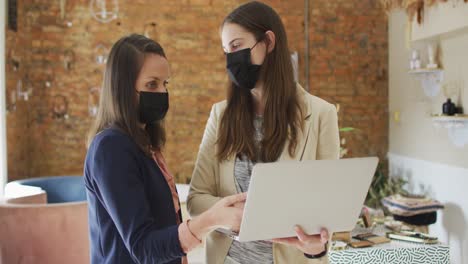 Caucasian-female-business-owner-and-her-coworker-wearing-face-masks,-using-laptop-and-talking