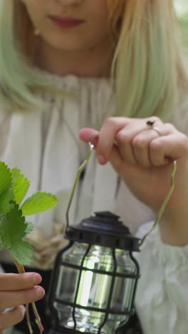 teenage girl in vintage dress looks at green leaves holding lamp with friends on wood glade closeup. medieval camp for children. role playing