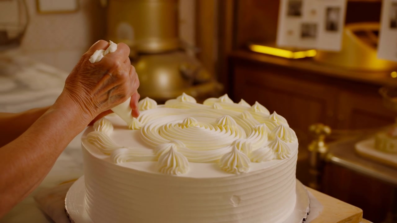 Scatto ravvicinato in movimento di mani mature che stringono una sac à poche per creare creste a spirale concentriche e rosette di crema al burro in rilievo sulla parte superiore di una torta a strati rotonda con luce calda da cucina, con torta, crema al burro