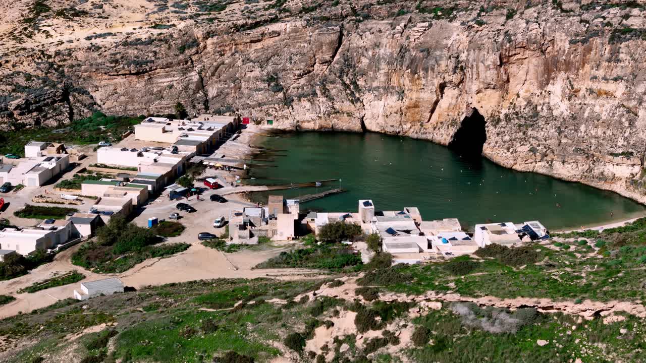 Góc nhìn từ trên cao của Biển nội địa Dwejra ở Gozo Malta dưới bầu trời mùa hè quang đãng, ánh nắng phản chiếu trên làn nước xanh lục và những vách đá vôi cổ bao bọc những nhà thuyền đánh cá mộc mạc và các hang động biển