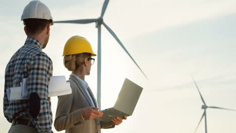 Side-view-of-Caucasian-woman-and-man-engineers-wearing-a-helmet-using-laptop-while-talking-about-operation-of-the-windmill-tourbines