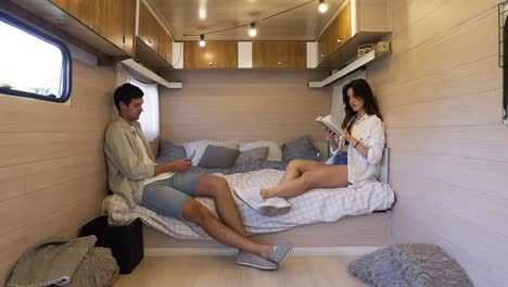 life in the trailer. joint evening together sitting on the bed - attractive, long haired woman reading a book while her husband scrolling phone. stylish, modern interior