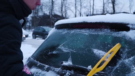 el joven está limpiando la ventana trasera del auto de la nieve con un cepillo amarillo en invierno