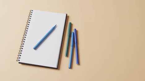 Close-up-of-blue-markers-with-notebook-with-copy-space-on-beige-background,-in-slow-motion