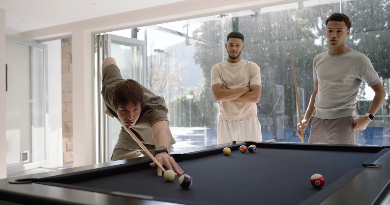 Diverse male friends watching shooter lining up, striking cue ball on black felt table for fun