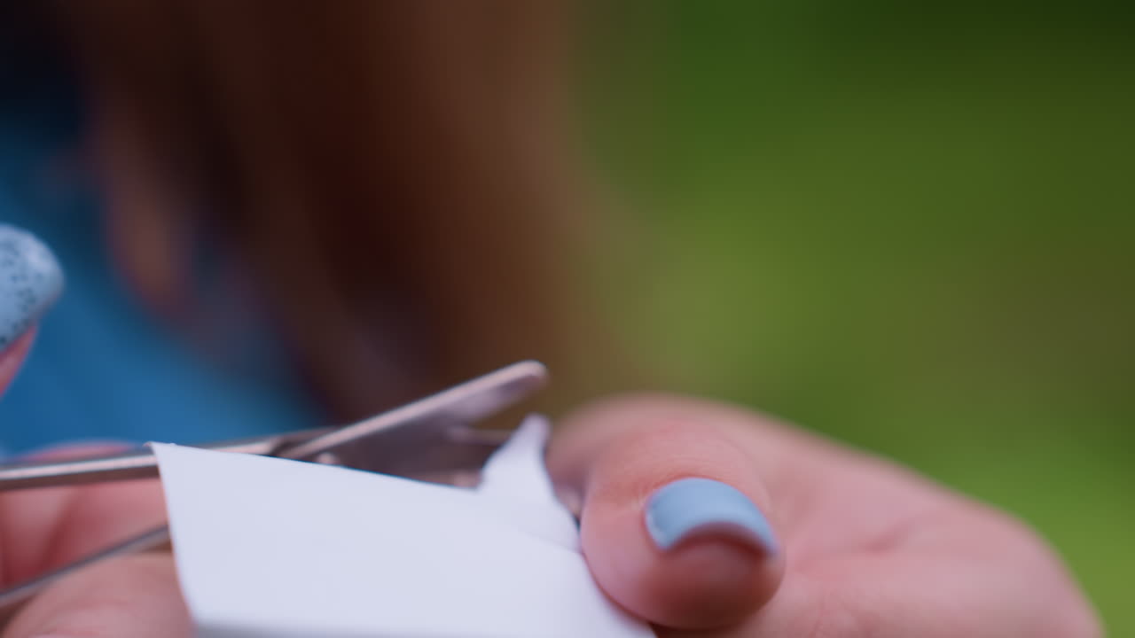 Close up hand view of lady with blue polished nails using small scissors to cut braided paper thread outdoors, showing careful precision and soft natural daylight with blurred greenery background