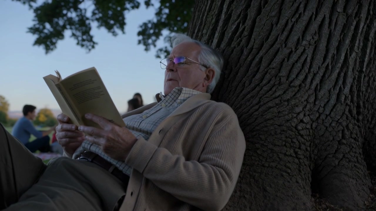 Reading senior in cardigan turning pages, camera pulling back against park tree, relaxing with book