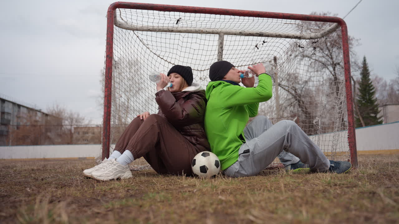 Белые подростки пьют воду, стоя спиной к спине у футбольных ворот, отдыхая после матча, пластиковые бутылки и общий смех, прерывистое дыхание, пятна травы на кроссовках, пригородный пейзаж на заднем плане, радостное восстановление сил