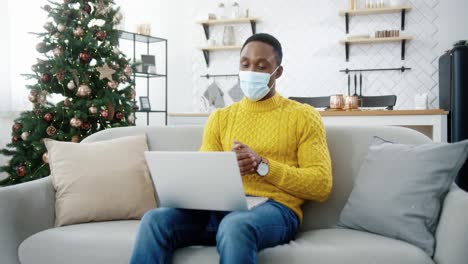 Chico-Guapo-Con-Máscara-Médica-Sentado-En-El-Sofá-En-Una-Habitación-Decorada-En-Casa-Cerca-Del-árbol-De-Navidad-Con-Luces-Centelleantes-Y-Chat-De-Video-En-Una-Laptop