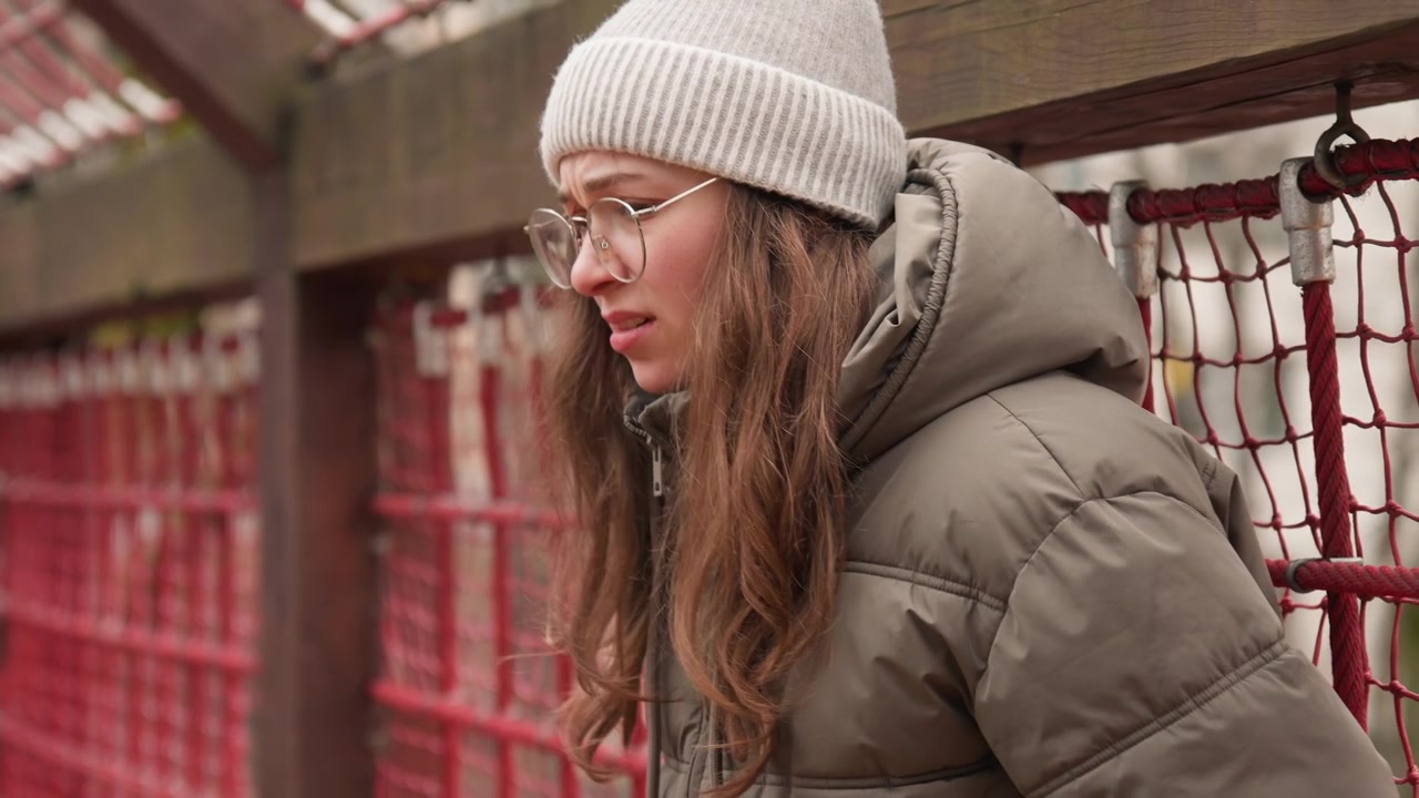 White woman on snowy footbridge, university student pausing between classes, leaning against red netting and wooden railing, wearing knit beanie and puffer jacket with round glasses, soft overcast