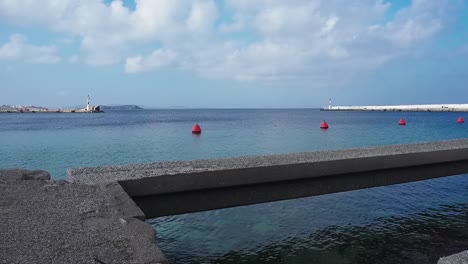 static view of a concrete bar hovering over shallow water with red buoys floating in the background of the old port of mykonos