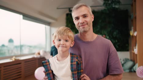 Portrait-of-a-Happy-male-teacher-with-gray-hair-and-stubble-in-a-purple-T-shirt-who-poses-with-his-student,-a-blond-boy-with-blue-eyes-in-a-checkered-shirt-and-a-white-T-shirt-against-the-background-of-other-children-who-are-having-fun-at-recess-in-a-club-for-preparing-for-school