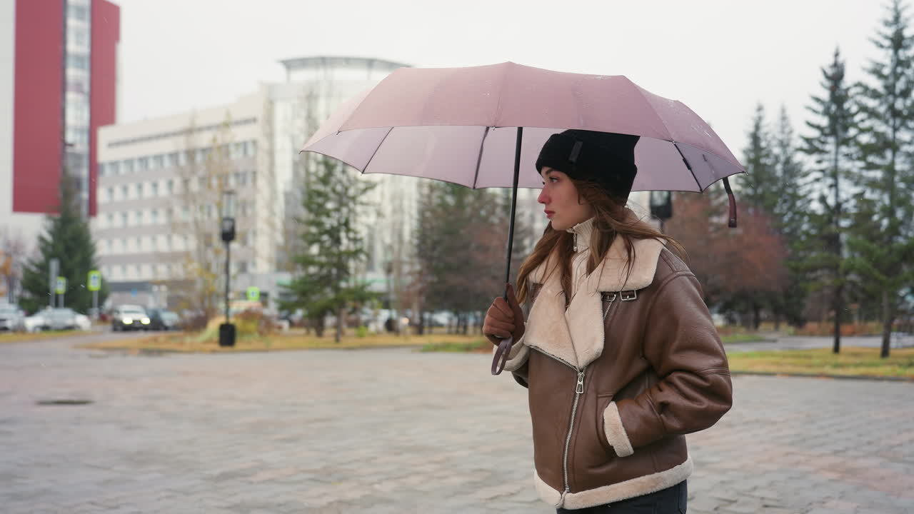 Thoughtful girl holding umbrella wearing black knit cap and brown shearling jacket, surrounded by pine trees and urban buildings, with soft snowfall falling on cold overcast day