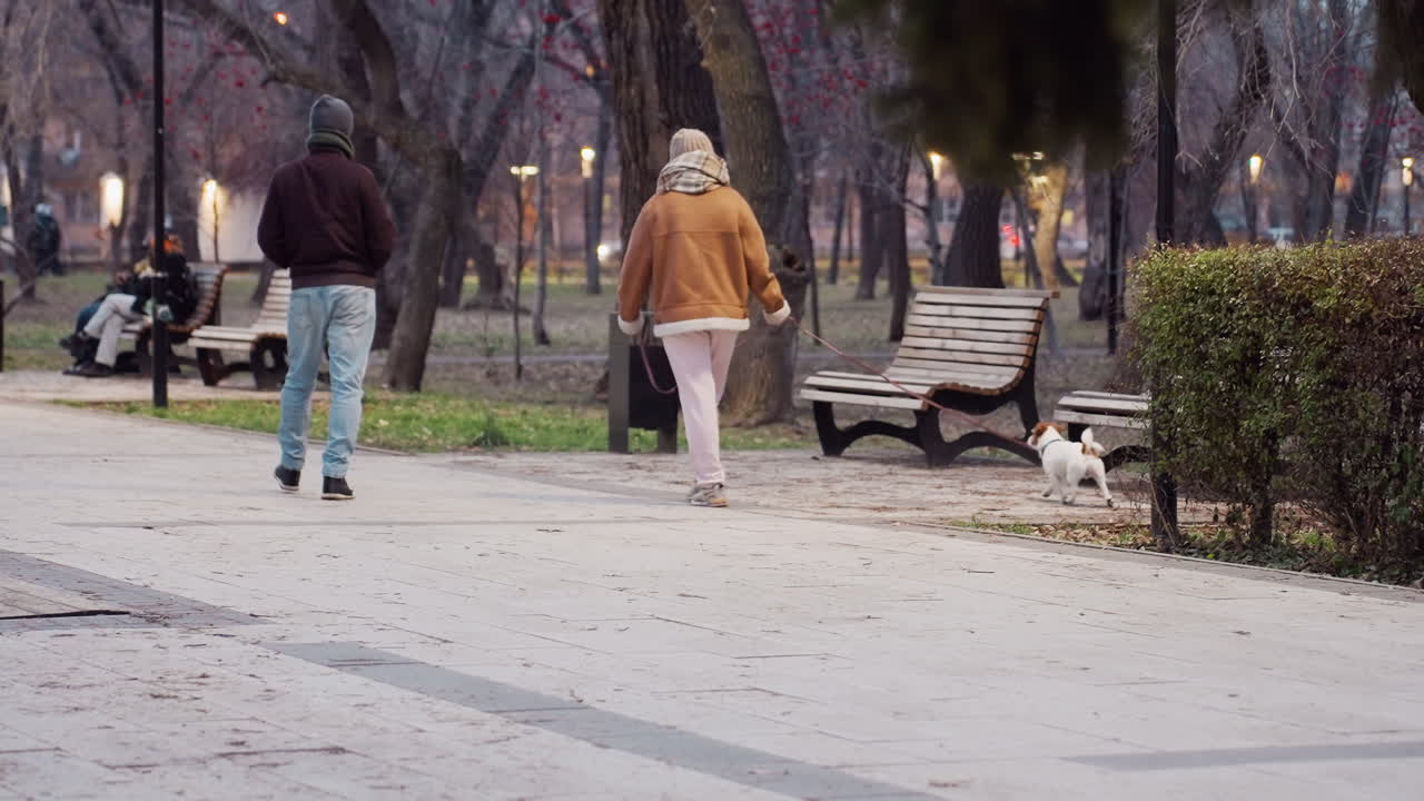 Recreational park scene with woman dressed in winter outfit walking small dog on leash beside man in warm clothes, benches and people visible in soft evening light atmosphere