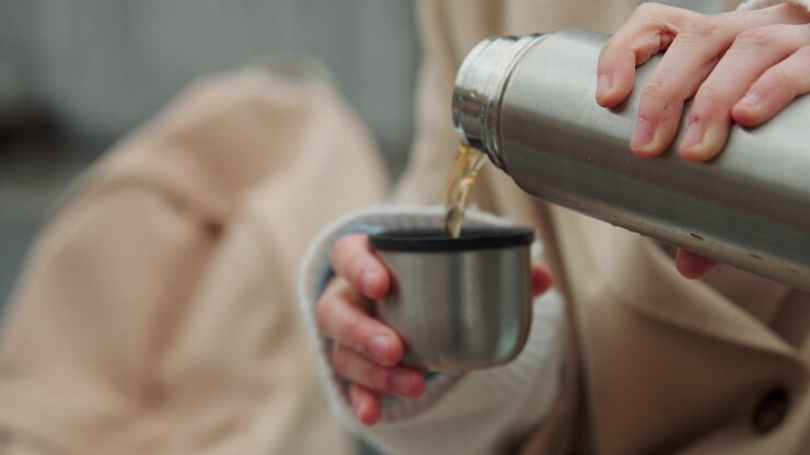 Primo piano di una persona che versa del liquido da un thermos in acciaio inossidabile in una tazza abbinata, con goccioline d'acqua visibili sulle superfici metalliche. La persona indossa maglieria di colore chiaro e tiene entrambi gli oggetti.
