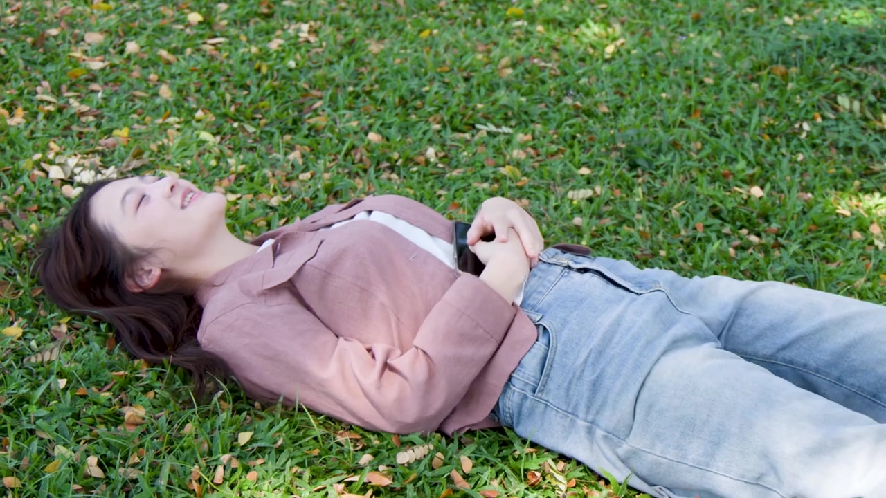 A young Asian woman adjusts her hair before lying down on grass in bright sunlight