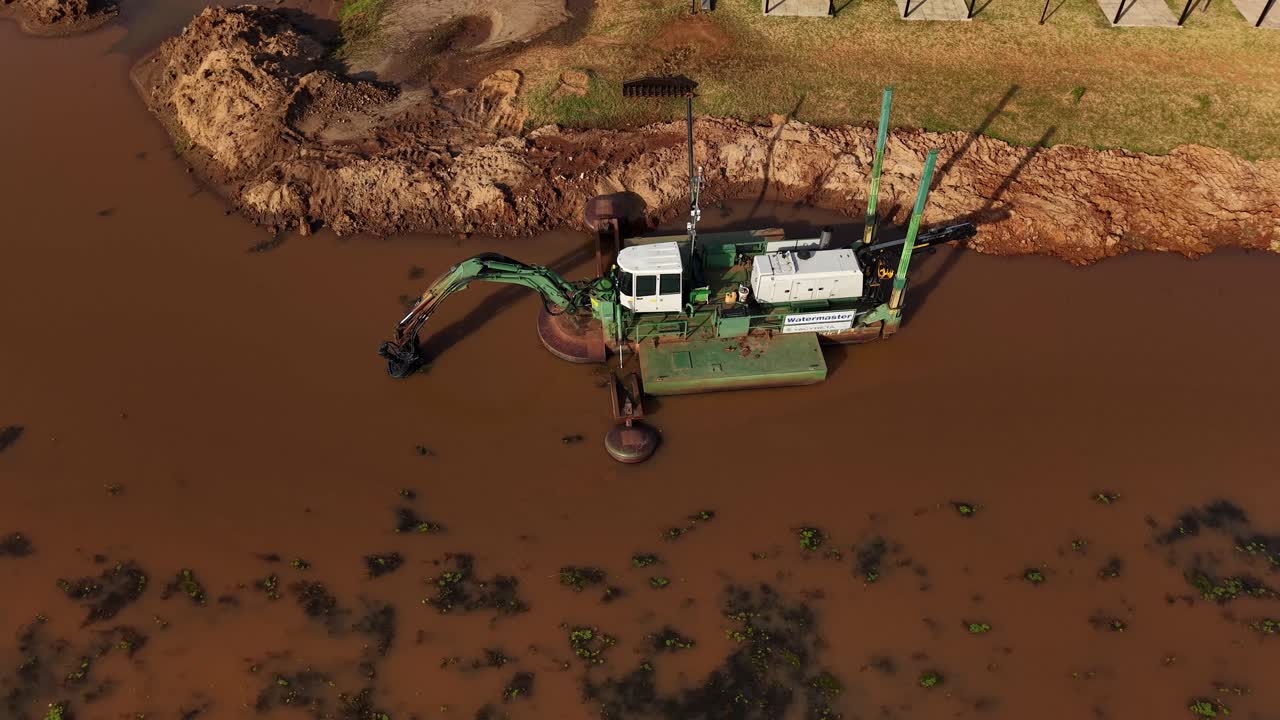 Aerial orbit over amphibious dredging machine operating on muddy river shoreline during afternoon with intense sun rays