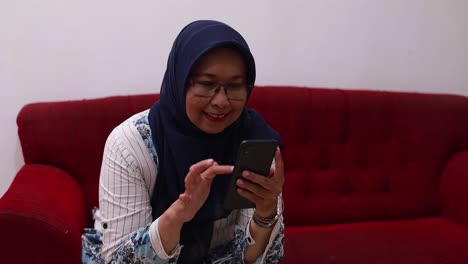 Happy-smiling-asian-elderly-woman-sitting-on-the-sofa-at-home-while-using-a-cell-phone