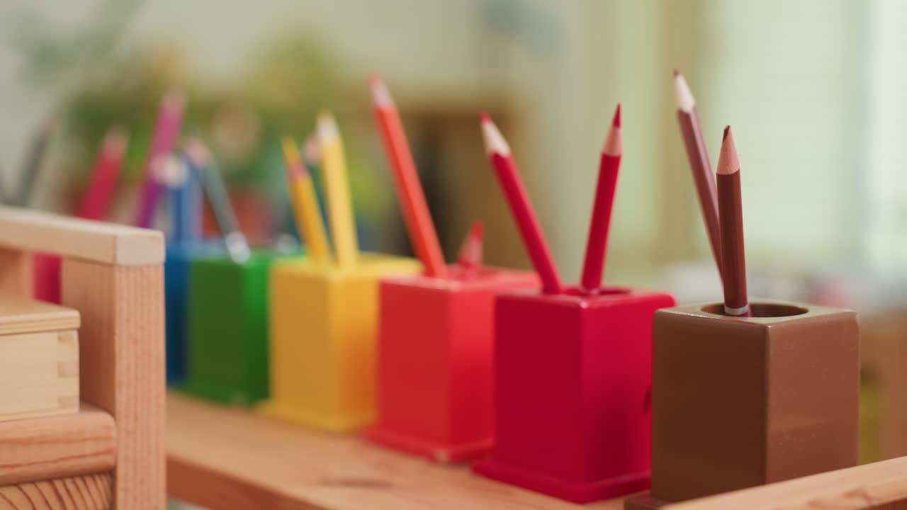 Close up of colored pencils in bright holders on wooden shelf, foreground crisp with blurred classroom background, showing creativity, learning, art supplies, tidy order, warm preschool environment
