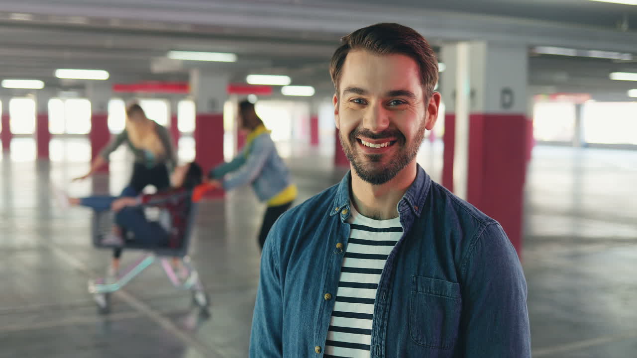 Portrait of Caucasian man standing in front of the camera and smiling while on the background his friends having fun with trolley in the parking
